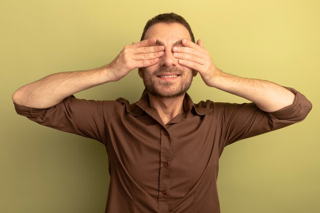 Улыбающийся молодой кавказский мужчина закрывает глаза руками, изолированными на оливково-зеленом фоне