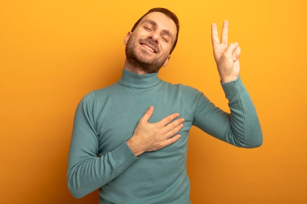 Smiling young caucasian man doing peace sign putting hand on chest with closed eyes isolated on orange wall with copy space