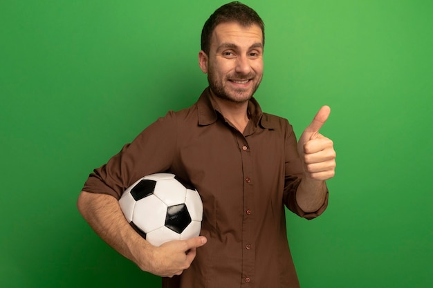 Sorridente giovane uomo caucasico tenendo il pallone da calcio guardando la telecamera che mostra il pollice in alto isolato su sfondo verde con copia spazio