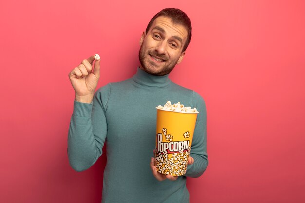 Smiling young caucasian man looking at camera holding bucket of popcorn and popcorn piece isolated on crimson background with copy space