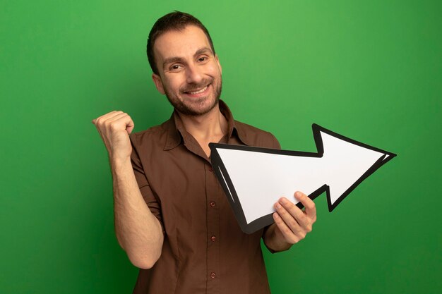 Smiling young caucasian man looking at camera pointing behind and holding arrow mark that is pointing at side isolated on green background with copy space