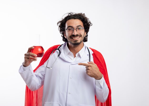 Smiling young caucasian man in optical glasses wearing doctor uniform with red cloak and stethoscope around neck holds and points at red chemical liquid in glass flask