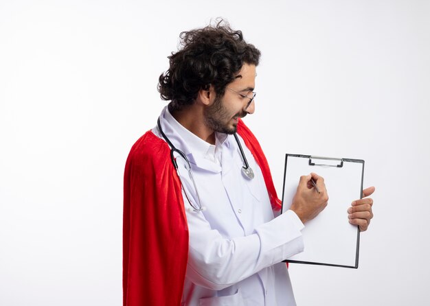 Smiling young caucasian man in optical glasses wearing doctor uniform with red cloak and with stethoscope around neck looks and writes on clipboard with pencil with copy space