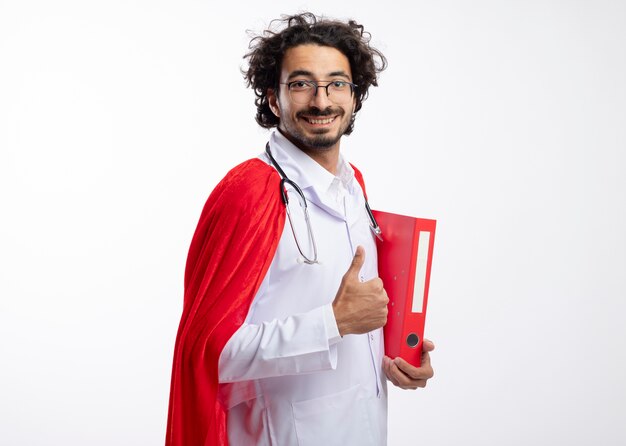 Smiling young caucasian man in optical glasses wearing doctor uniform with red cloak and with stethoscope around neck stands sideways holding file folder and thumbs up with copy space