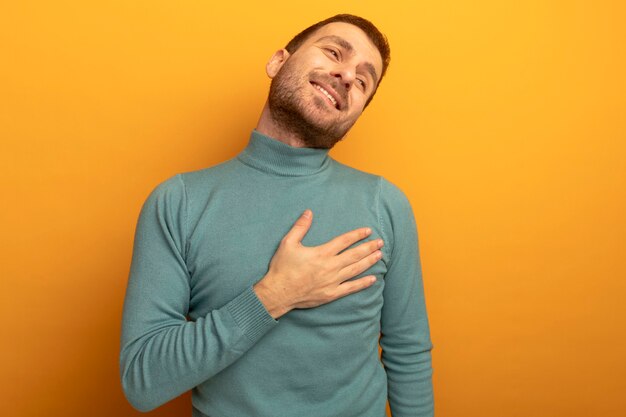 Smiling young caucasian man putting hand on chest looking at side isolated on orange wall with copy space