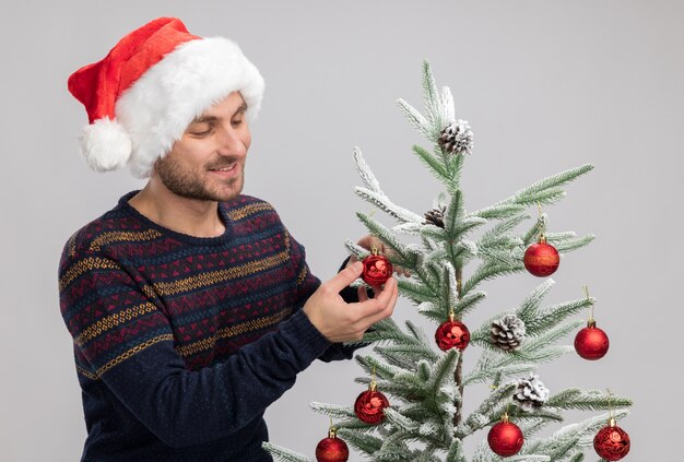 улыбающийся молодой кавказец в новогодней шапке стоит возле елки и смотрит на нее трогательно рождественская безделушка, изолированная на белой стене