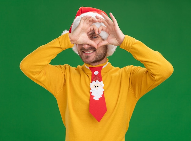 Smiling young caucasian man wearing christmas hat and tie looking at camera doing heart sign in front of eye isolated on green background