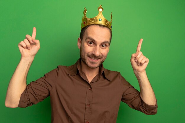 Smiling young caucasian man wearing crown  pointing up isolated on green wall