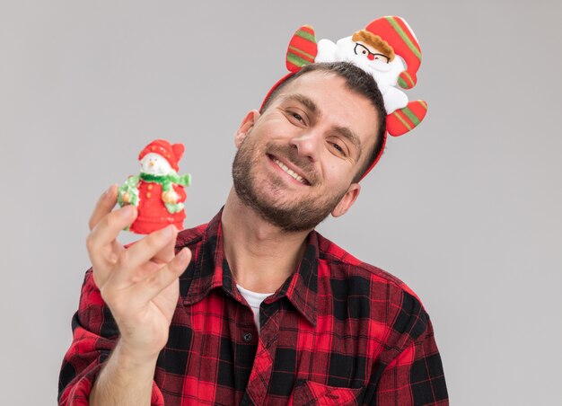 Smiling young caucasian man wearing santa claus headband holding snowman christmas ornament looking at camera isolated on white background