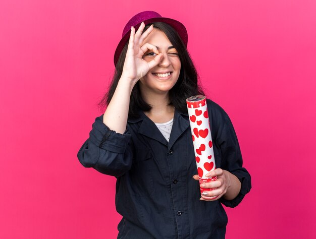 Улыбающаяся молодая кавказская тусовщица в партийной шляпе держит конфетти-пушку и делает жест, изолированный на розовой стене