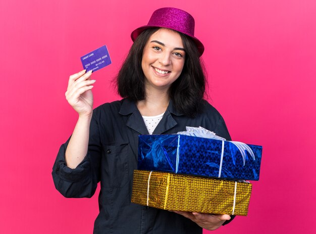 Улыбающаяся молодая кавказская тусовщица в партийной шляпе держит подарочные пакеты и кредитную карту, изолированную на розовой стене