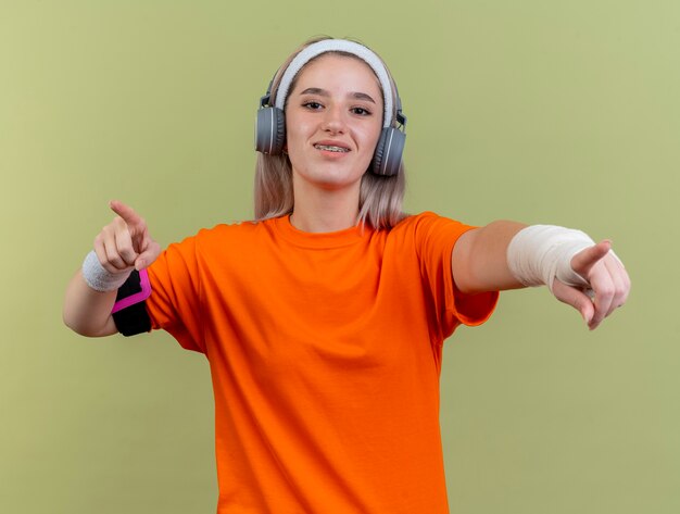 Улыбающаяся молодая кавказская спортивная девушка с подтяжками на наушниках с повязкой на голову и браслетами и повязкой для телефона указывает на камеру двумя руками