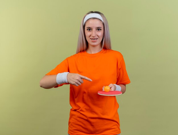 Smiling young caucasian sporty girl with braces wearing headband and wristbands holds and points at ping-pong ball on racket 