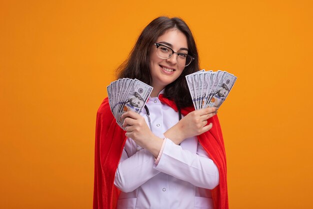 Smiling young caucasian superhero girl wearing doctor uniform and stethoscope with glasses keeping hands crossed holding money 