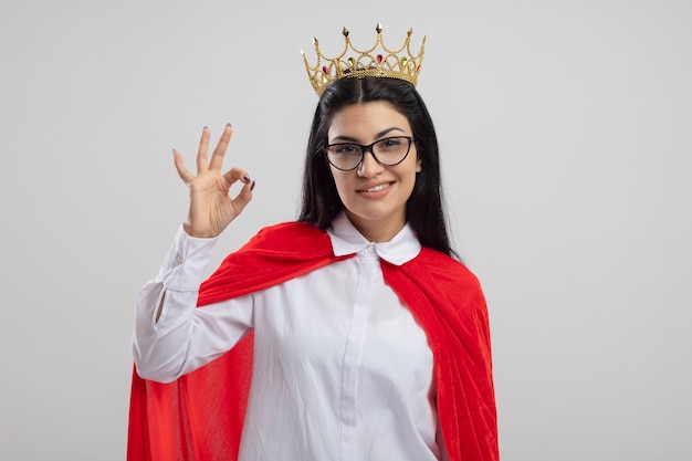 Smiling young caucasian superhero girl wearing glasses and crown looking at camera doing ok sign isolated on white background with copy space