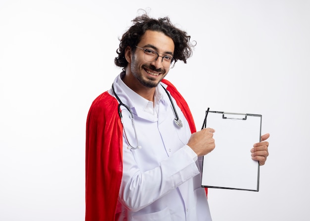 Smiling young caucasian superhero man in optical glasses wearing doctor uniform with red cloak and with stethoscope around neck points at clipboard holding pencil with copy space