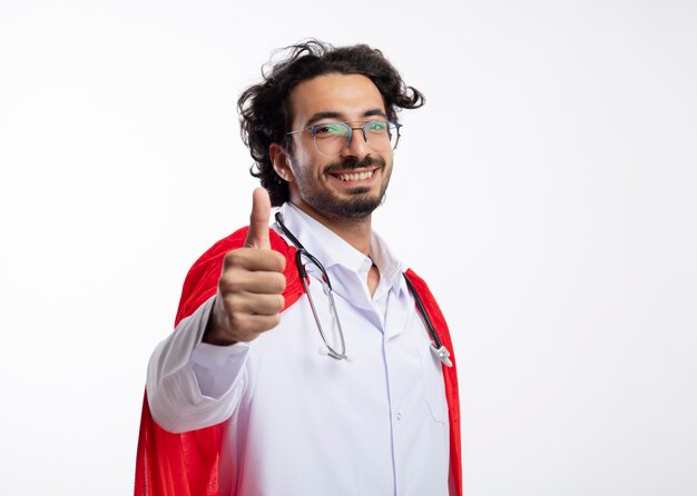 빨간 망토와 목 엄지 손가락 주위에 청진기와 의사 유니폼을 입고 광학 안경에 웃는 젊은 백인 슈퍼 히어로 남자