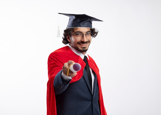 Smiling young caucasian superhero man in optical glasses wearing suit with red cloak and graduation cap holds out diploma 