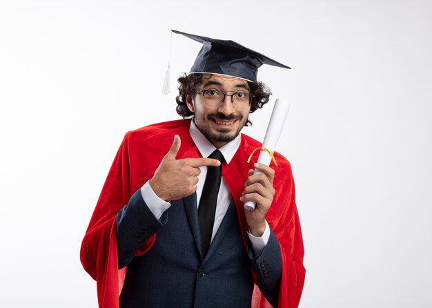 Smiling young caucasian superhero man in optical glasses wearing suit with red cloak and graduation cap holds and points at diploma 