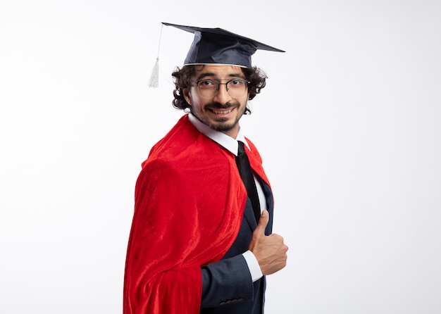 Free photo smiling young caucasian superhero man in optical glasses wearing suit with red cloak and graduation cap stands sideways and thumbs up