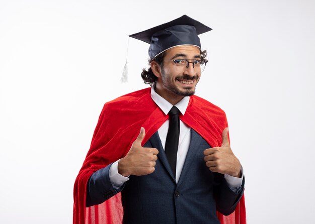 赤いマントと卒業帽のスーツを着て光学メガネで笑顔の若い白人のスーパーヒーローの男は、コピースペースと白い背景で隔離の両手で親指を立てる