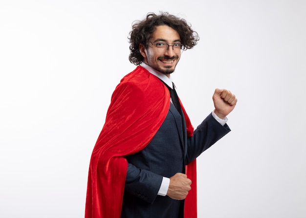 Smiling young caucasian superhero man in optical glasses wearing suit with red cloak stands sideways pretending to run isolated on white background with copy space