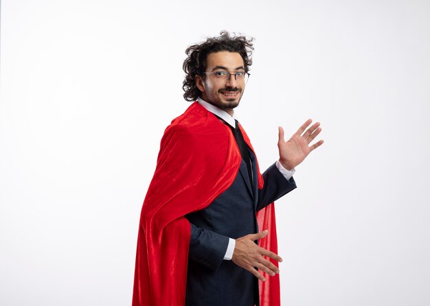 Smiling young caucasian superhero man in optical glasses wearing suit with red cloak stands sideways with raised hands looking at camera 