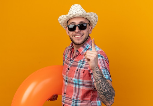 Sorridente giovane viaggiatore caucasico con cappello da spiaggia di paglia in occhiali da sole che tiene anello da nuoto e punta verso l'alto isolato sulla parete arancione con spazio per le copie