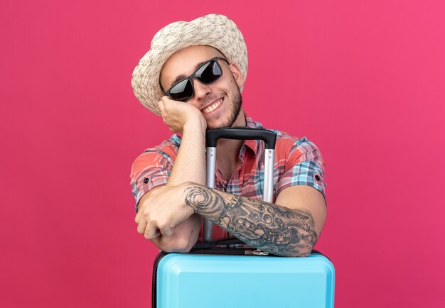 Smiling young caucasian traveler man with straw beach hat in sun glasses putting hands on suitcase isolated on pink background with copy space