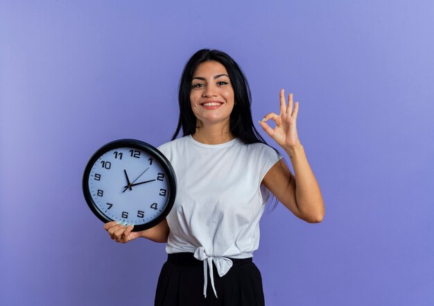 Smiling young caucasian woman gestures ok hand sign and holds clock