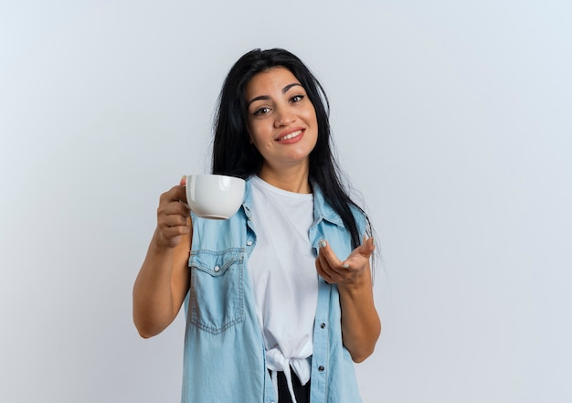 Smiling young caucasian woman holds cup and points at camera