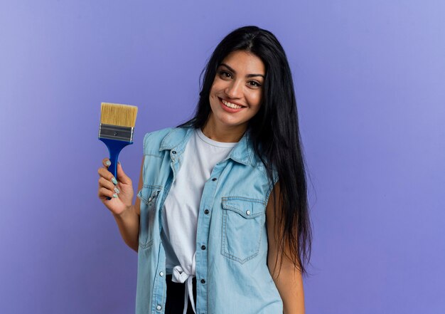 Smiling young caucasian woman holds paint brush looking at camera isolated on purple background with copy space