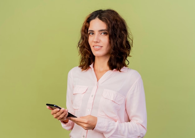 La giovane donna caucasica sorridente tiene il telefono che guarda l'obbiettivo isolato su priorità bassa verde con lo spazio della copia