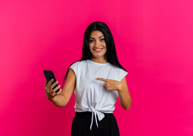 Smiling young caucasian woman holds and points at phone