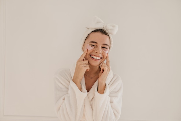 무료 사진 테리 목욕 가운을 입은 웃고 있는 젊은 백인 여성이 눈을 가늘게 뜨고 흰색 배경에 투명한 패치를 적용합니다. 홈 스파 페이셜 컨셉