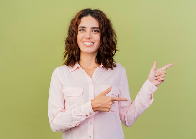 Smiling young caucasian woman points at side with two hands isolated on green background with copy space