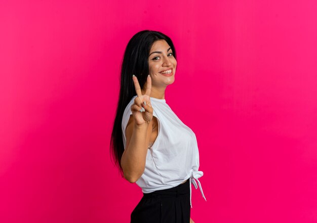 Smiling young caucasian woman stands sideways gesturing victory hand sign