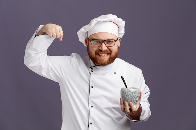 Smiling young chef wearing glasses uniform and cap holding bowl with spoon in it looking at camera showing strong gesture isolated on purple background