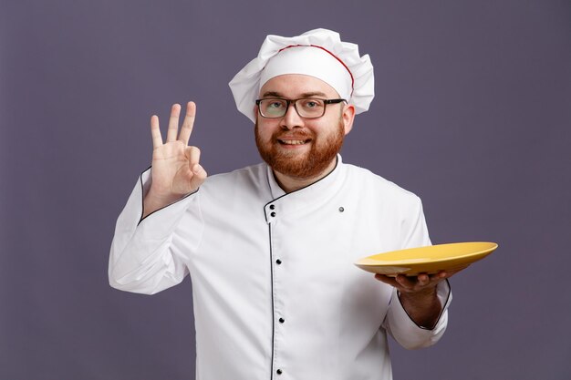 Sorridente giovane chef con gli occhiali uniforme e cappuccio che tiene piatto vuoto guardando la fotocamera che mostra segno ok isolato su sfondo viola