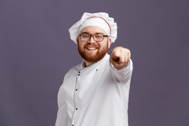 Улыбающийся молодой шеф-повар в очках и кепке смотрит и указывает на камеру, изолированную на фиолетовом фоне