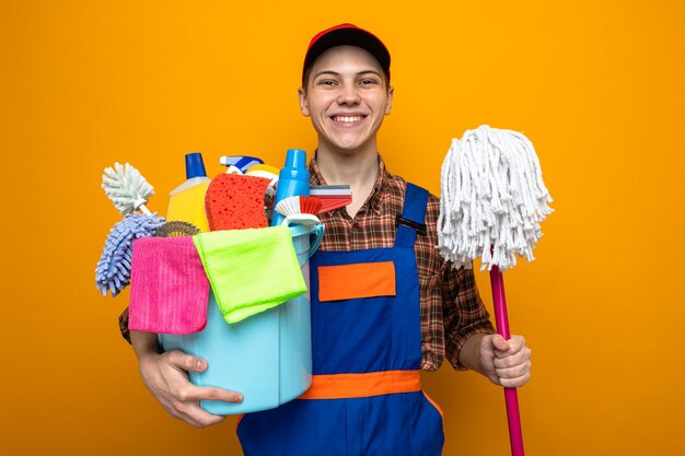 Smiling young cleaning guy wearing uniform and cap holding bucket of cleaning tools with mop 