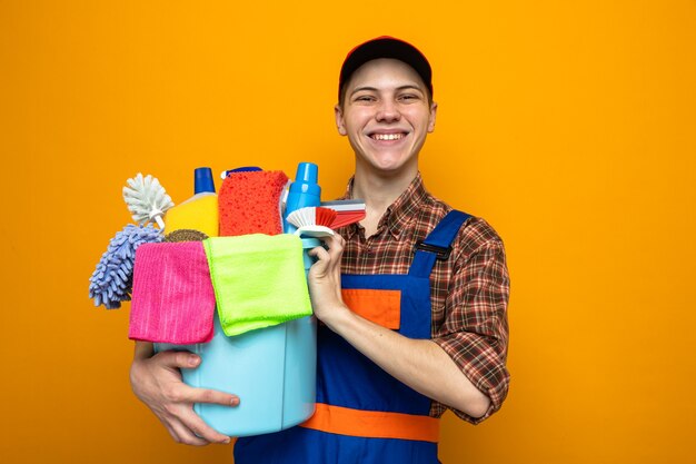Smiling young cleaning guy wearing uniform and cap holding bucket of cleaning tools 