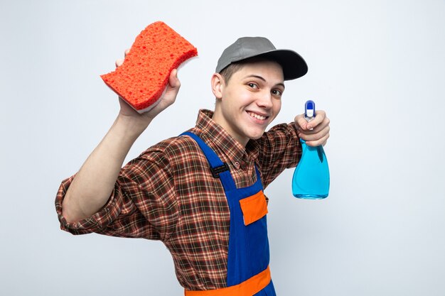 Smiling young cleaning guy wearing uniform and cap holding sponge with cleaning agent 