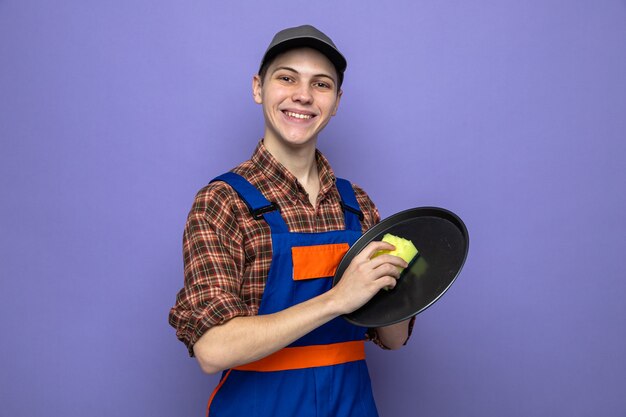 Smiling young cleaning guy wearing uniform and cap washing tray with sponge 