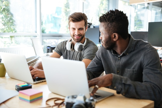 Smiling young colleagues sitting in office coworking using laptop