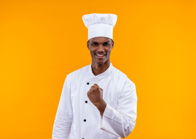 Smiling young confident afro-american cook in chef uniform keeps fist up isolated on orange background with copy space