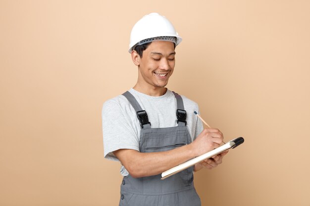 Улыбающийся молодой строитель в защитном шлеме и форме письма карандашом на блокноте