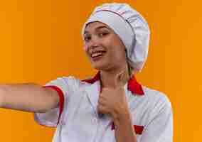 Free photo smiling young cook female wearing chef uniform holding out camera her thumb up on isolated orange background