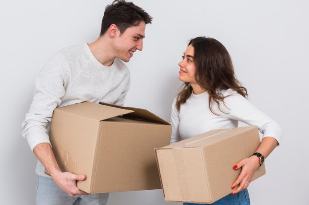 Smiling young couple carrying cardboard boxes in hand looking at each other against white background