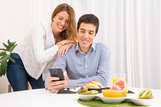 Улыбается молодая пара, глядя на мобильный телефон за столом для завтрака